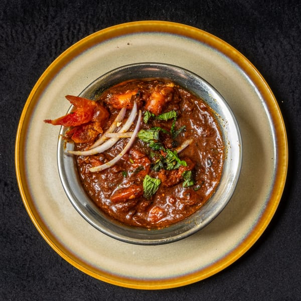 Anjappar Chettinad Indian Restaurant's chicken curry, garnished with mint and onions, in a metal bowl.