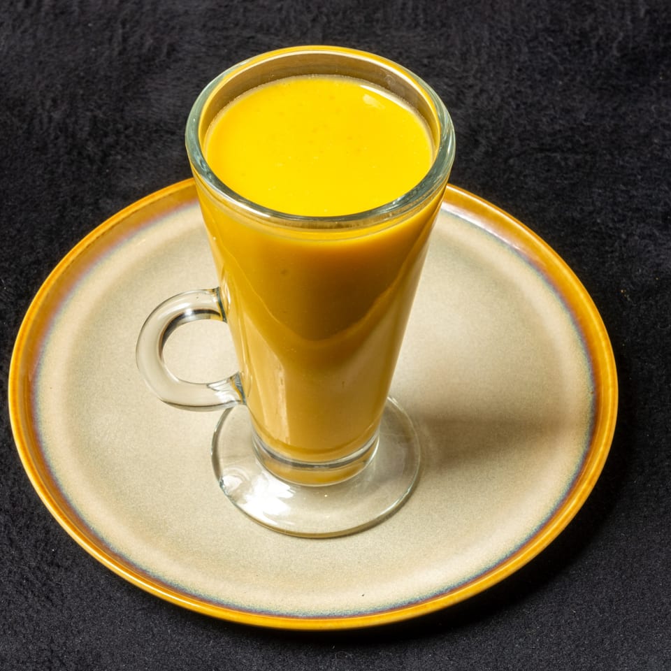 A tall glass of bright orange mango lassi sits on a beige and orange plate.