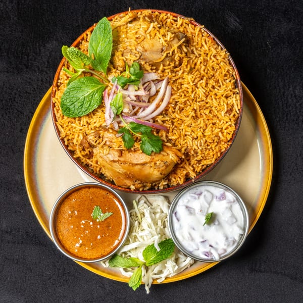 Anjappar Chicken Biryani with rice, chicken, mint, onions, and two side sauces on a yellow plate.