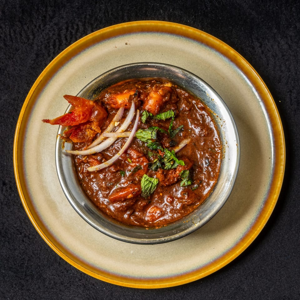 Anjappar Chettinad Indian Restaurant's chicken curry, garnished with onions and mint, in a metal bowl.