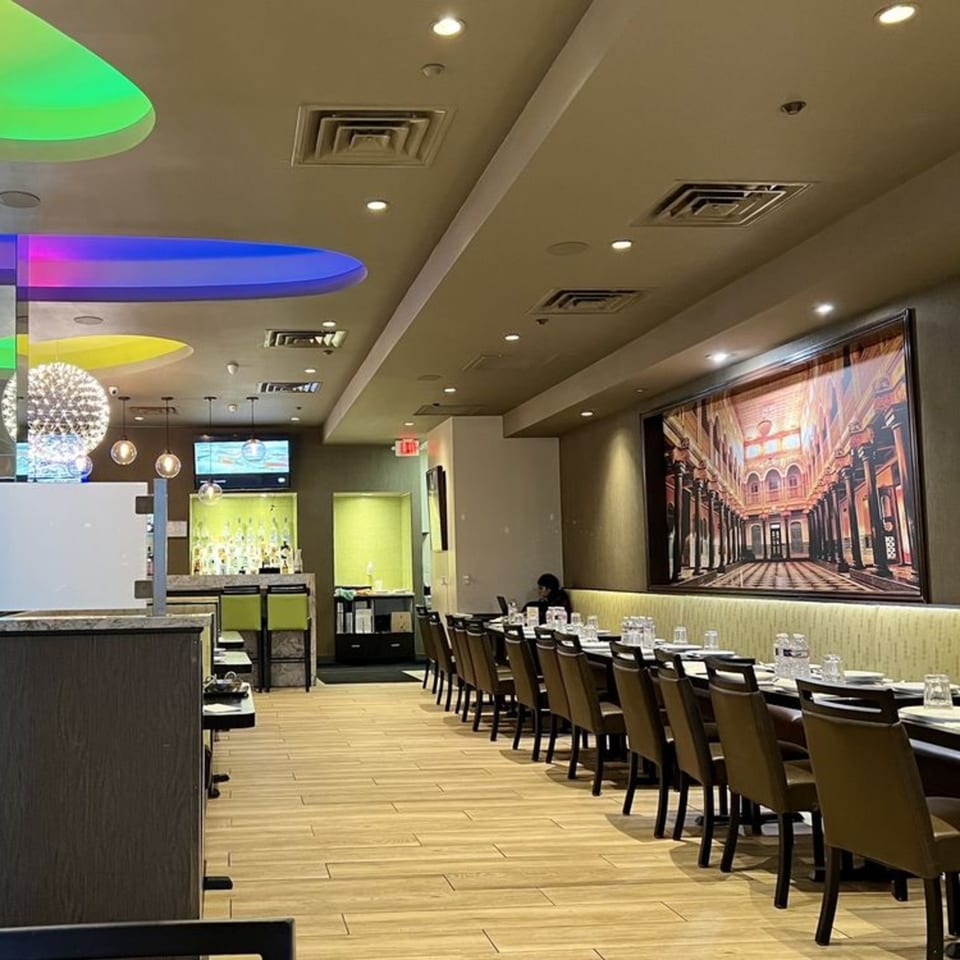 Anjappar Chettinad Indian Restaurant interior with long dining tables, a bar, and colorful ceiling lights.